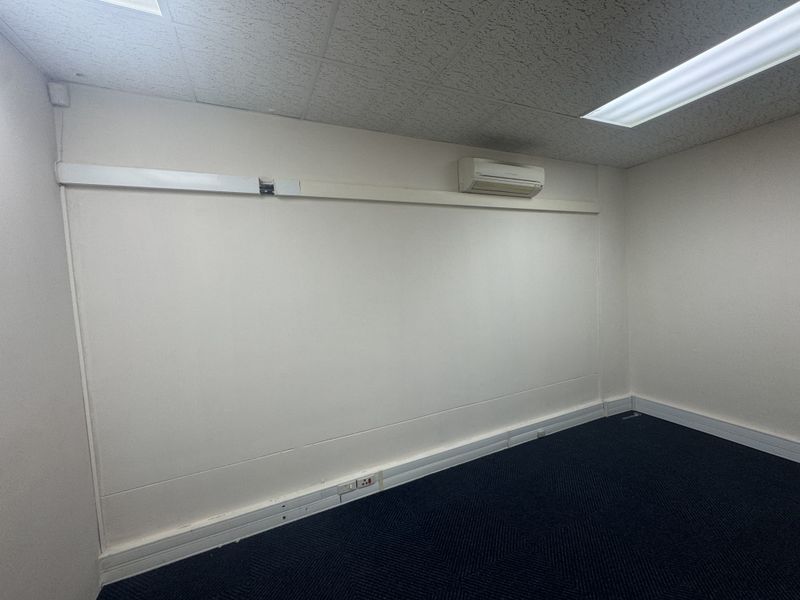 An empty office space featuring a suspended ceiling, recessed lighting, wall-mounted air conditioning, and blue carpeted flooring.