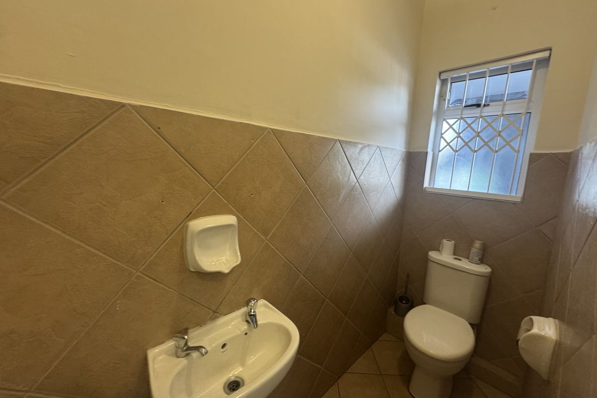 A compact office restroom featuring a pedestal washbasin, toilet, and diamond-patterned ceramic wall tiling with a security-barred window.