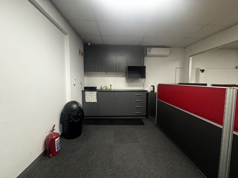 A functional kitchenette area featuring grey built-in cabinetry, a stainless steel sink, and a wall-mounted air conditioning unit within an office environment.