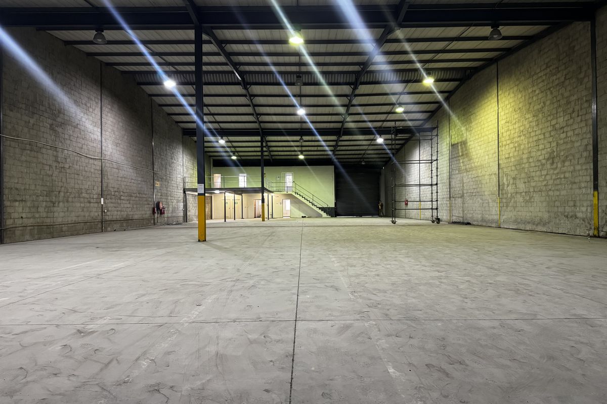 Spacious industrial warehouse featuring high ceilings, exposed steel structure, concrete flooring, a mezzanine level with staircase, and a large roller shutter door.
