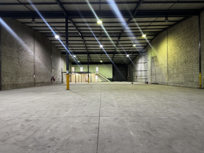 Spacious industrial warehouse featuring high ceilings, exposed steel structure, concrete flooring, a mezzanine level with staircase, and a large roller shutter door.