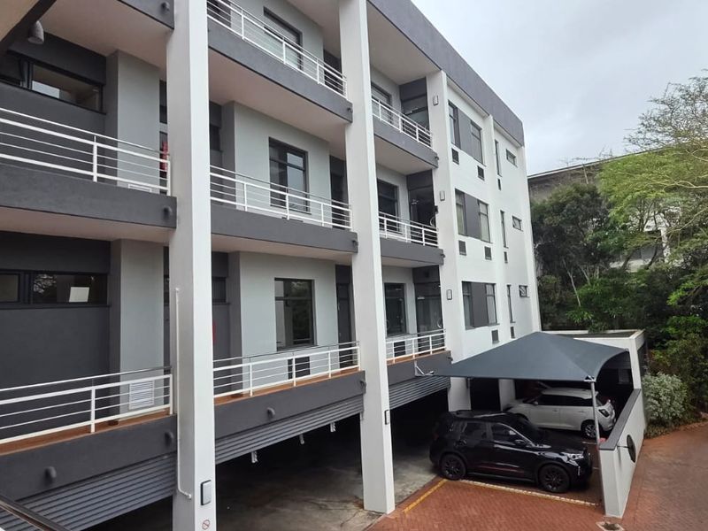 A modern multi-story office building facade featuring balconies with metal railings, covered parking bays, and a paved driveway entrance.