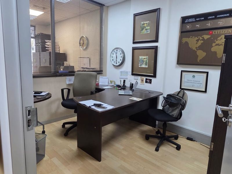 A functional office space featuring laminate flooring, an internal glass partition with blinds, and recessed ceiling lighting.