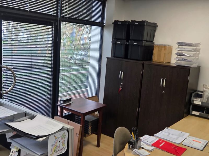 A functional office space featuring large windows with venetian blinds and laminate flooring, providing a bright and practical work environment.