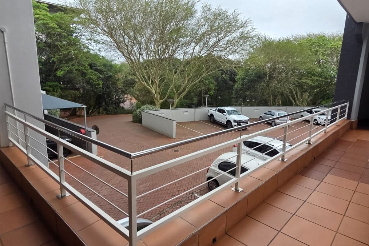 An elevated outdoor balcony area with tiled flooring and a metal balustrade, overlooking a paved parking area and surrounding greenery.