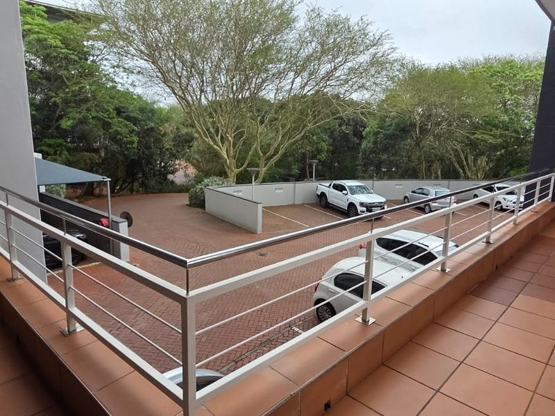 An elevated outdoor balcony area with tiled flooring and a metal balustrade, overlooking a paved parking area and surrounding greenery.
