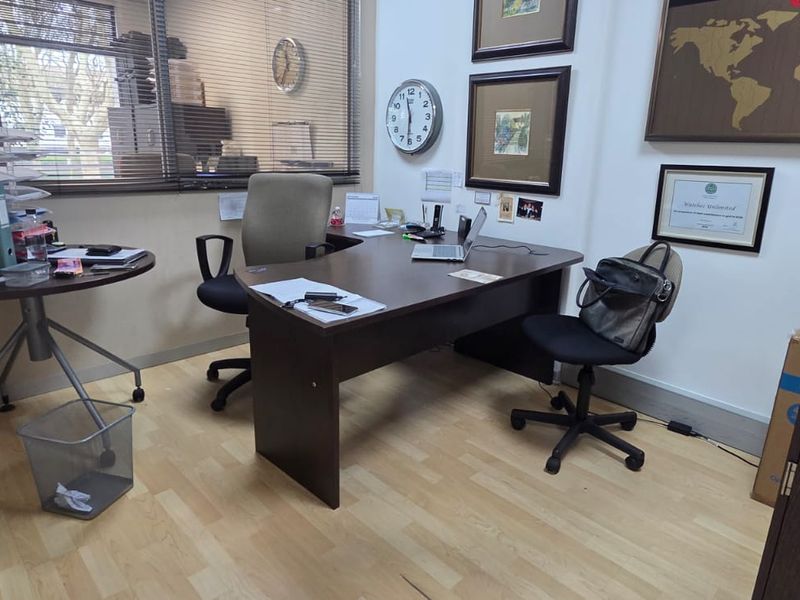 A functional office space featuring light wood-look laminate flooring and large windows with venetian blinds, providing a practical workspace environment.