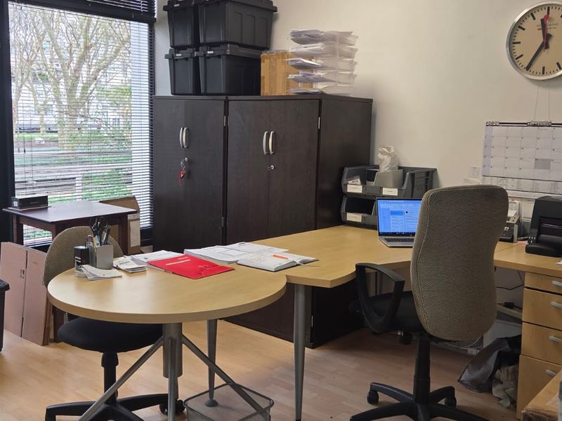 A functional office space featuring large windows with venetian blinds and light-toned laminate flooring, providing a practical work environment.
