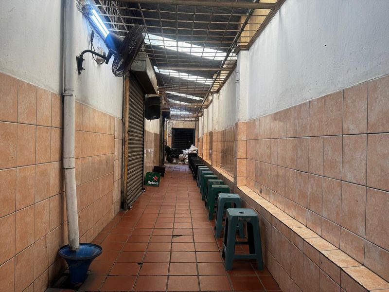 A narrow, covered outdoor walkway or patio area featuring tiled walls and flooring, with a corrugated roof and wall-mounted fan.