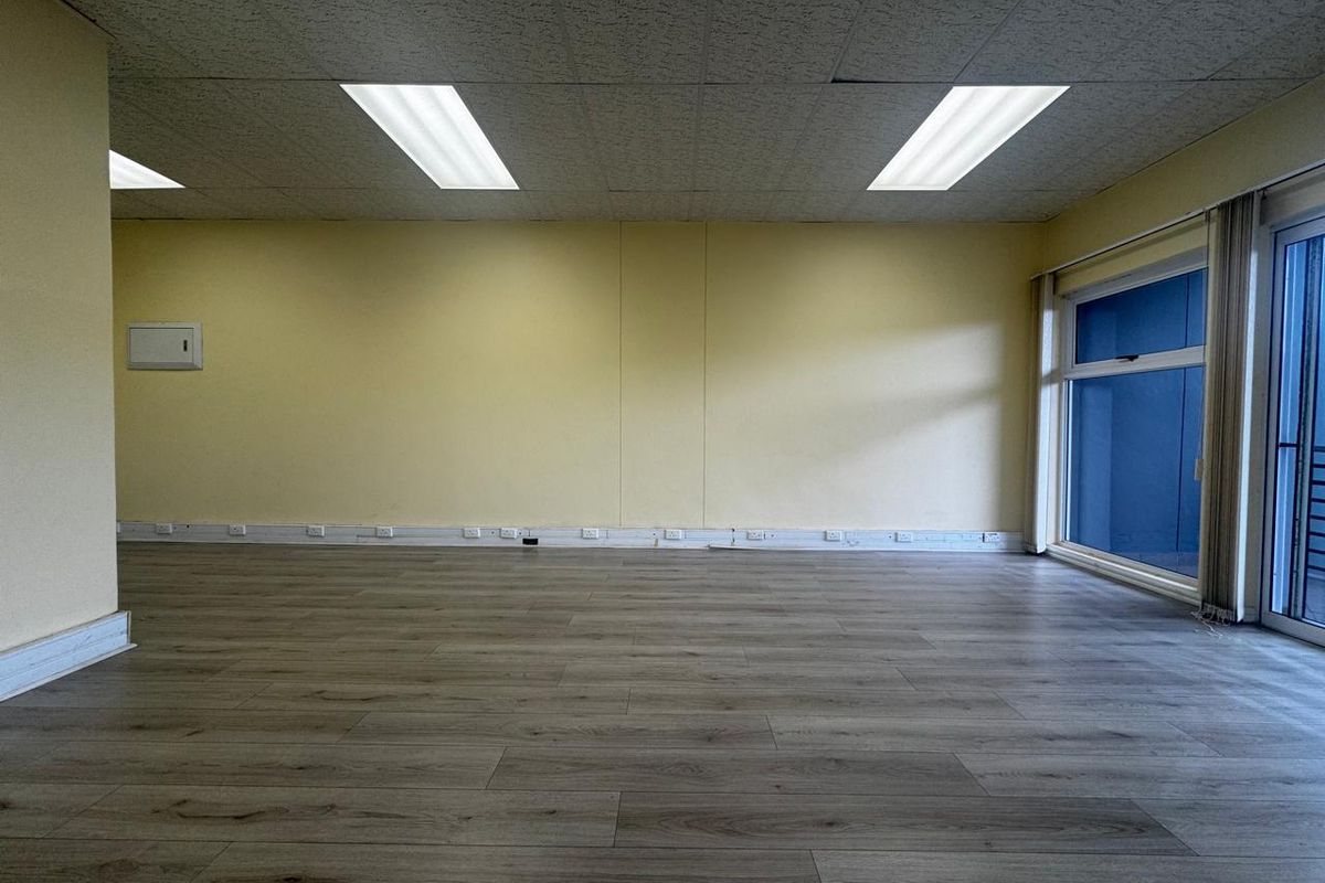 An empty, open-plan office space featuring laminate flooring, a suspended ceiling with recessed lighting, and large windows providing natural light.