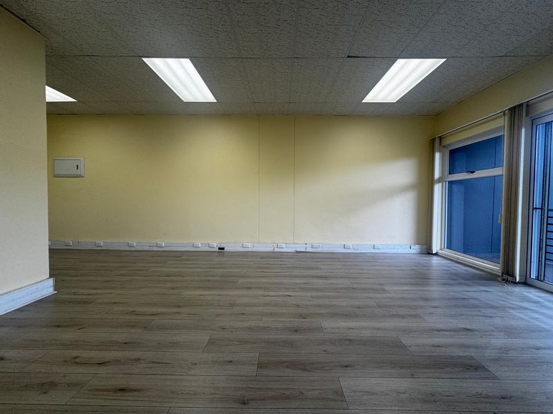 An empty, open-plan office space featuring laminate flooring, a suspended ceiling with recessed lighting, and large windows providing natural light.