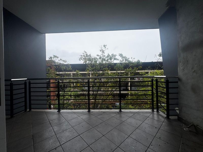 A covered balcony featuring tiled flooring and a secure metal balustrade, offering an elevated view of the surrounding greenery and neighboring buildings.
