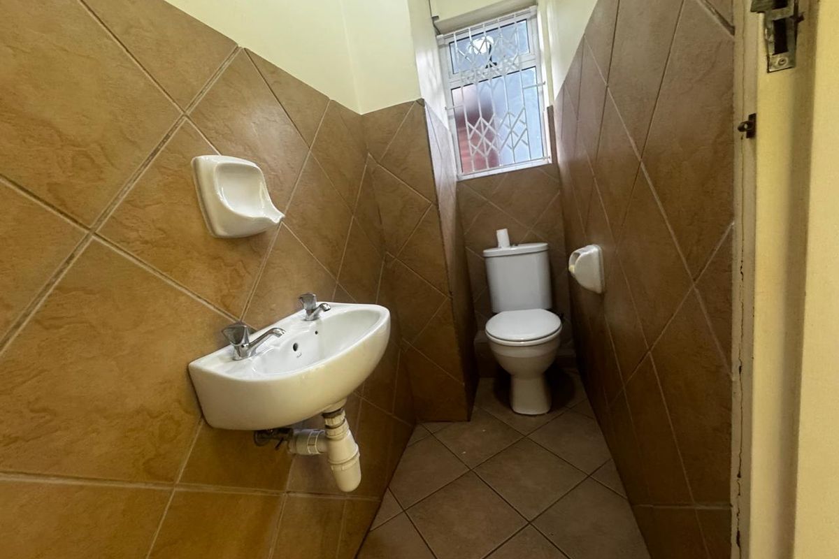 A compact office bathroom featuring a wall-mounted basin, toilet, and full-height ceramic wall tiling with a small window for ventilation.