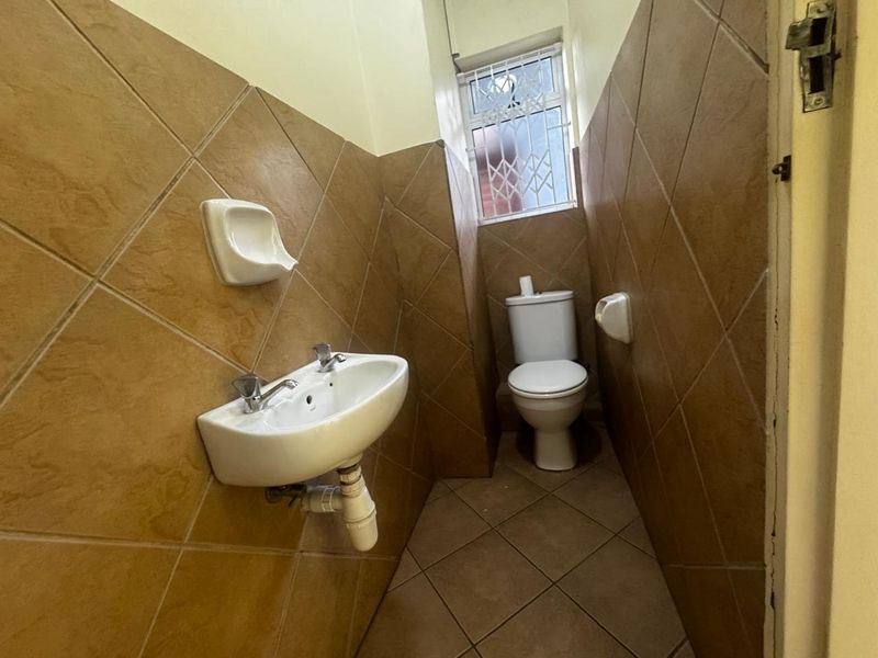A compact office bathroom featuring a wall-mounted basin, toilet, and full-height ceramic wall tiling with a small window for ventilation.