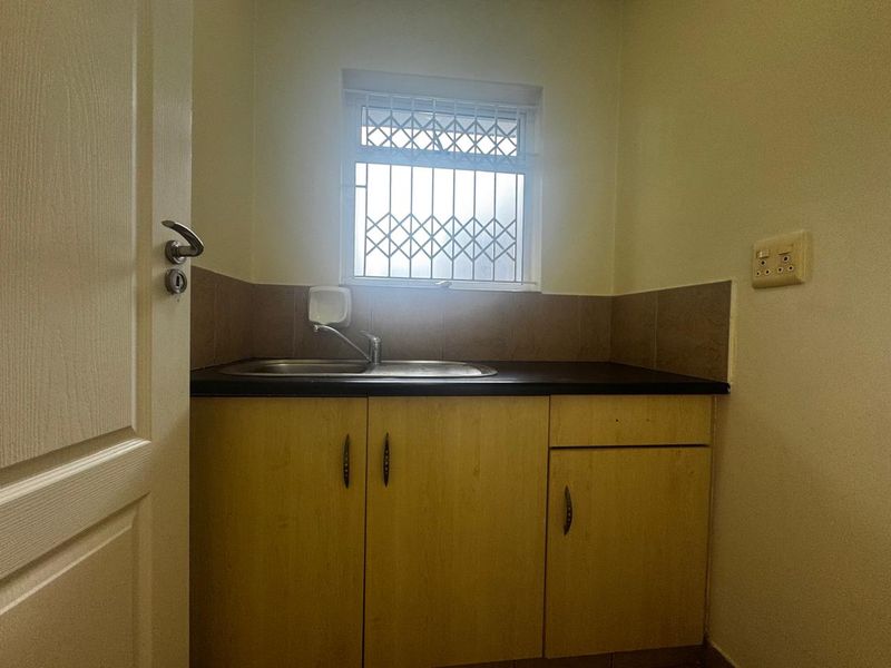 A compact scullery or utility area featuring built-in cabinetry, a stainless steel sink, and a tiled splashback under a window with security bars.