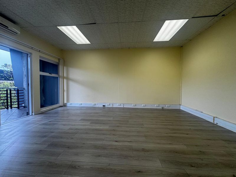 An empty office space featuring laminate flooring, a suspended ceiling with recessed lighting, wall-mounted air conditioning, and access to an outdoor balcony.