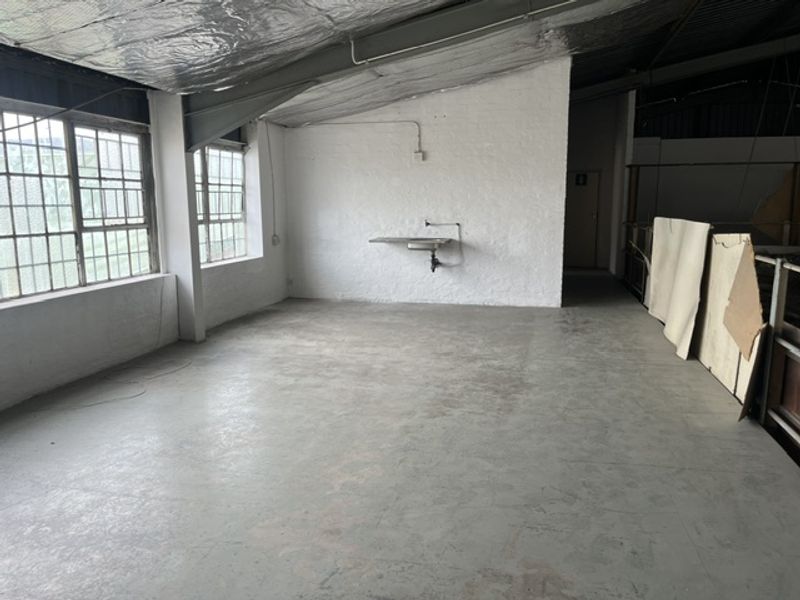 An open-plan industrial utility space featuring concrete flooring, exposed ceiling insulation, large steel-framed windows, and a wall-mounted utility sink.
