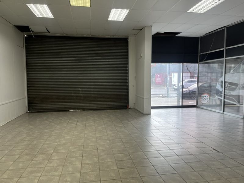 Empty industrial open-plan space featuring a large roller shutter door, tiled flooring, and a prominent glass storefront with street visibility.