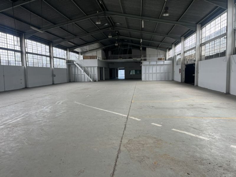 Spacious industrial warehouse featuring high ceilings with exposed steel trusses, concrete flooring, large multi-pane windows, and a mezzanine level with staircase access.