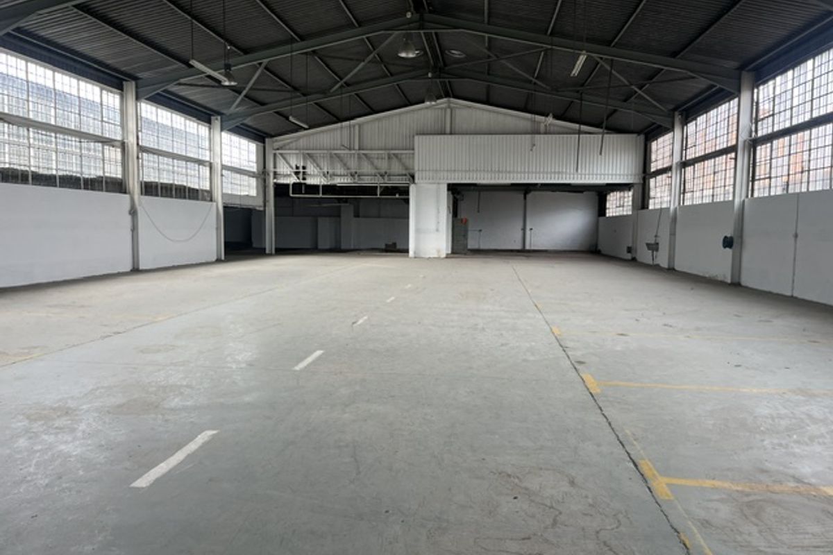 Spacious industrial warehouse featuring high ceilings with exposed steel trusses, concrete flooring, and large steel-framed windows providing ample natural light.