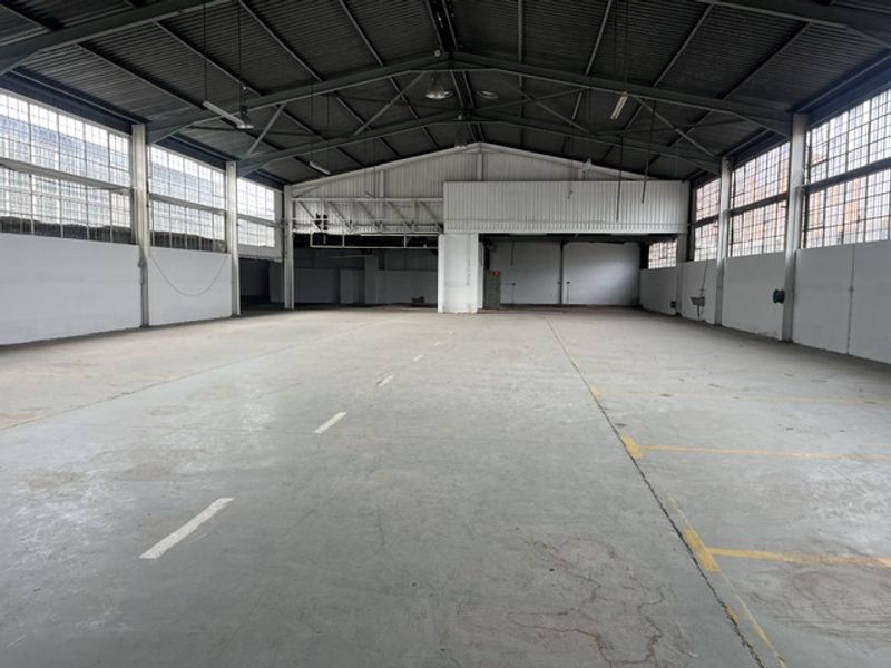Spacious industrial warehouse featuring high ceilings with exposed steel trusses, concrete flooring, and large steel-framed windows providing ample natural light.