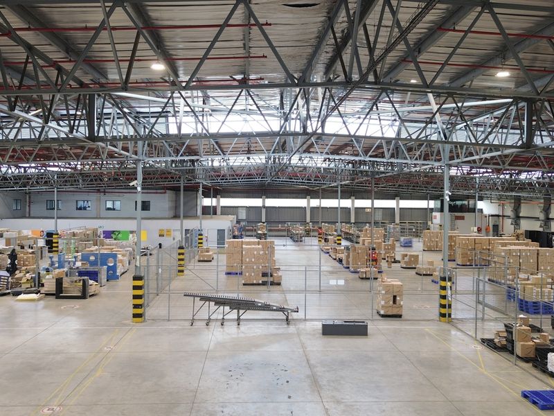 Large industrial warehouse space featuring high-clearance exposed steel truss roofing, concrete flooring, and integrated fire sprinkler systems with wire mesh partitioned storage areas.