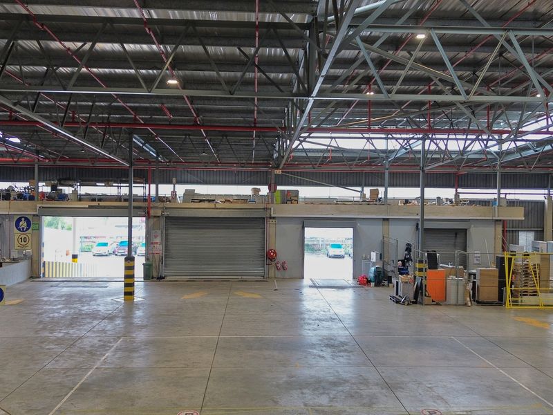 Spacious industrial warehouse featuring high ceilings with exposed steel trusses, concrete flooring, multiple roller shutter doors, and a functional mezzanine level.