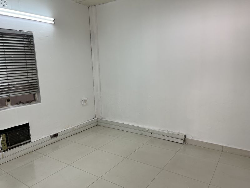 An empty office space featuring tiled flooring, a window-mounted air conditioning unit, and surface-mounted electrical trunking along the baseboards.