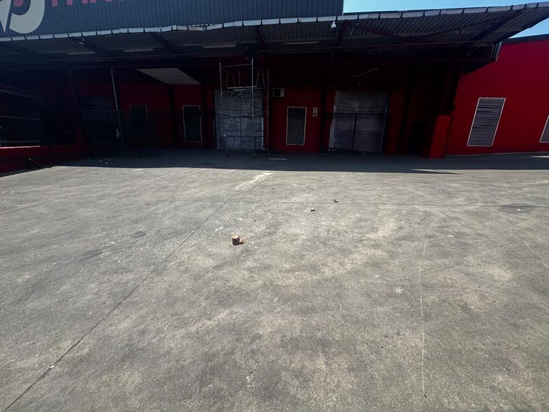 Industrial rear loading yard featuring a large concrete surface, multiple metal roller shutter doors, and a protective corrugated metal roof canopy.