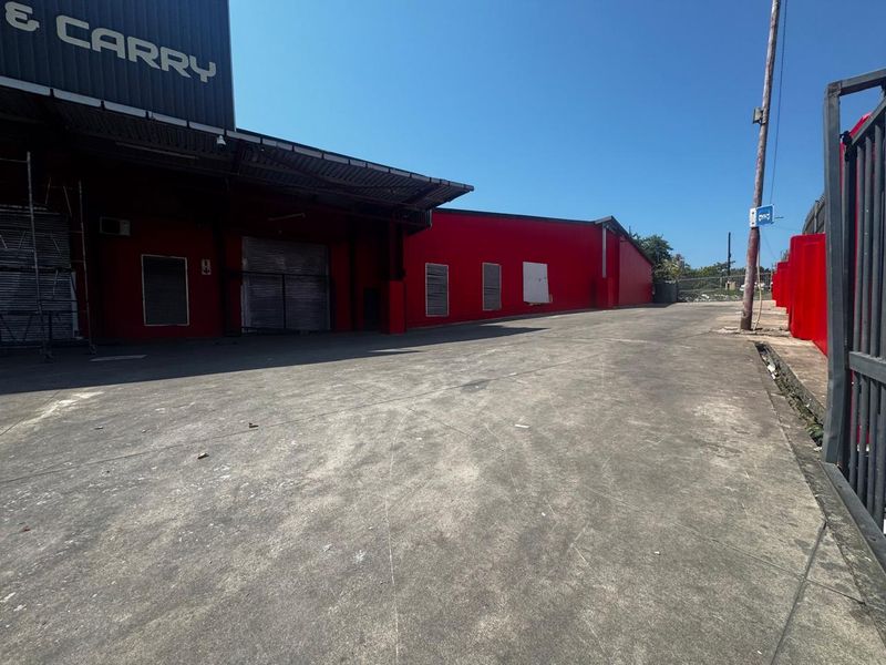 Exterior view of an industrial warehouse facility featuring a large concrete yard, metal cladding, and secure roller shutter access points.