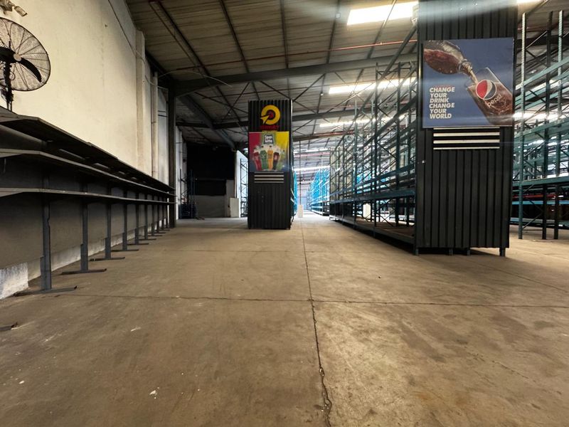 Spacious industrial warehouse area featuring high ceilings, exposed steel roof trusses, concrete flooring, and extensive built-in steel racking systems.