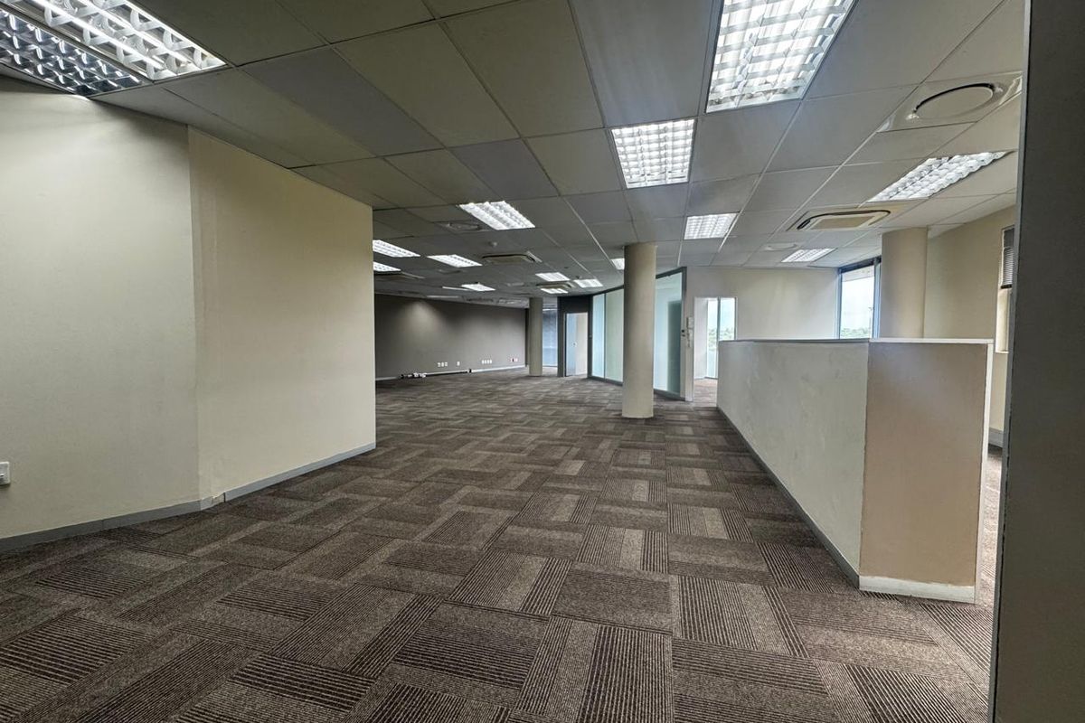 Spacious open-plan office area featuring suspended ceilings, recessed lighting, carpet tiles, and structural columns, offering a versatile workspace layout.
