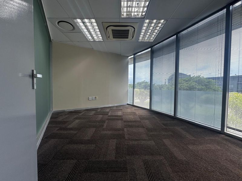 A bright, empty office space featuring floor-to-ceiling windows with venetian blinds, carpet tiles, and integrated cassette air conditioning.