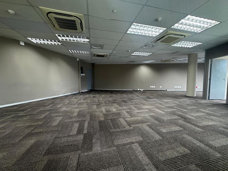 A spacious, open-plan office area featuring carpeted flooring, a suspended ceiling with integrated lighting, and cassette air conditioning units.