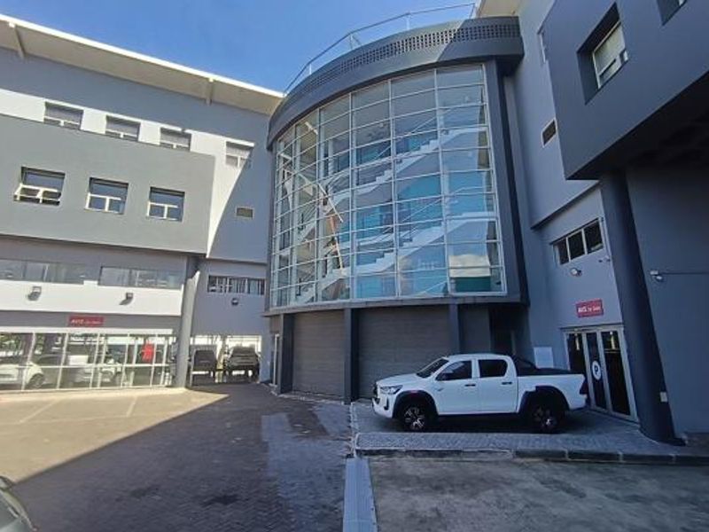 Modern commercial office building exterior featuring a prominent glass-enclosed stairwell, paved parking area, and contemporary grey facade finishes.