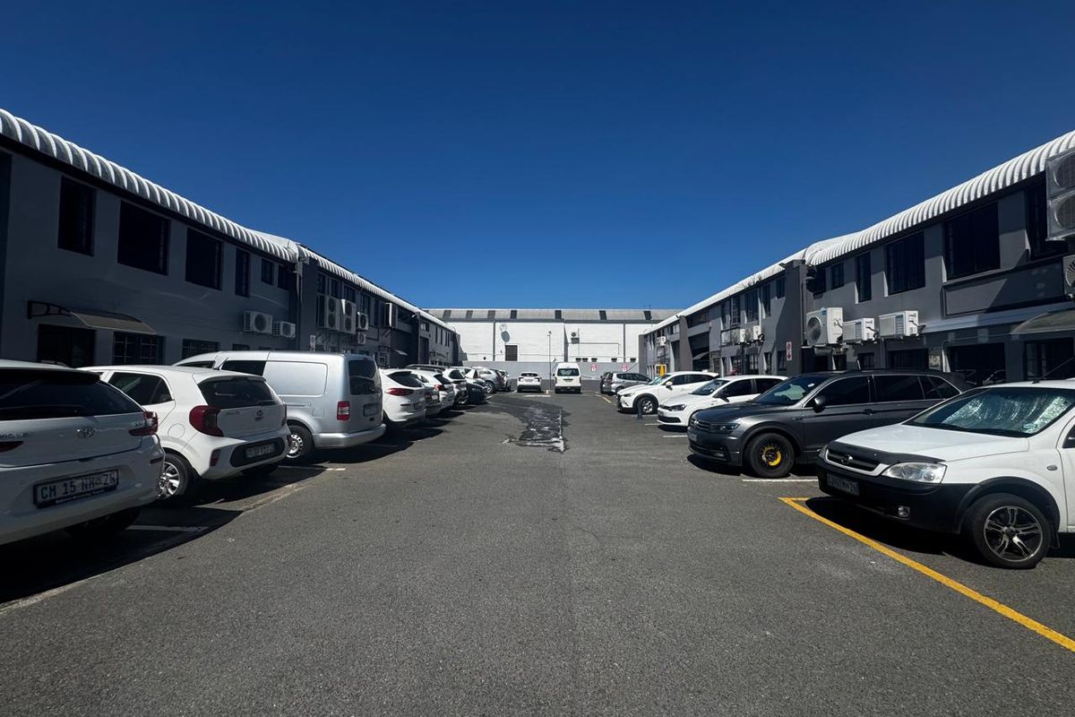 An industrial complex exterior featuring a paved parking area between two parallel warehouse buildings with mounted air conditioning units.