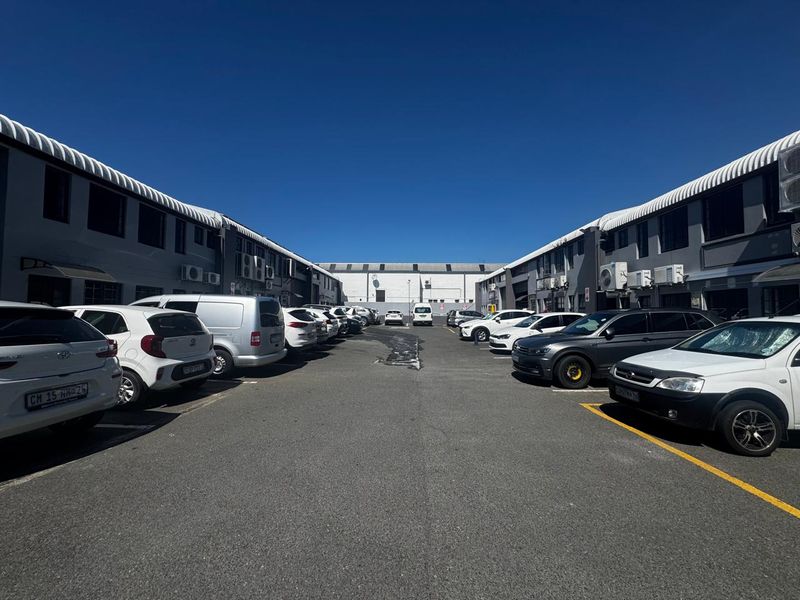 An industrial complex exterior featuring a paved parking area between two parallel warehouse buildings with mounted air conditioning units.