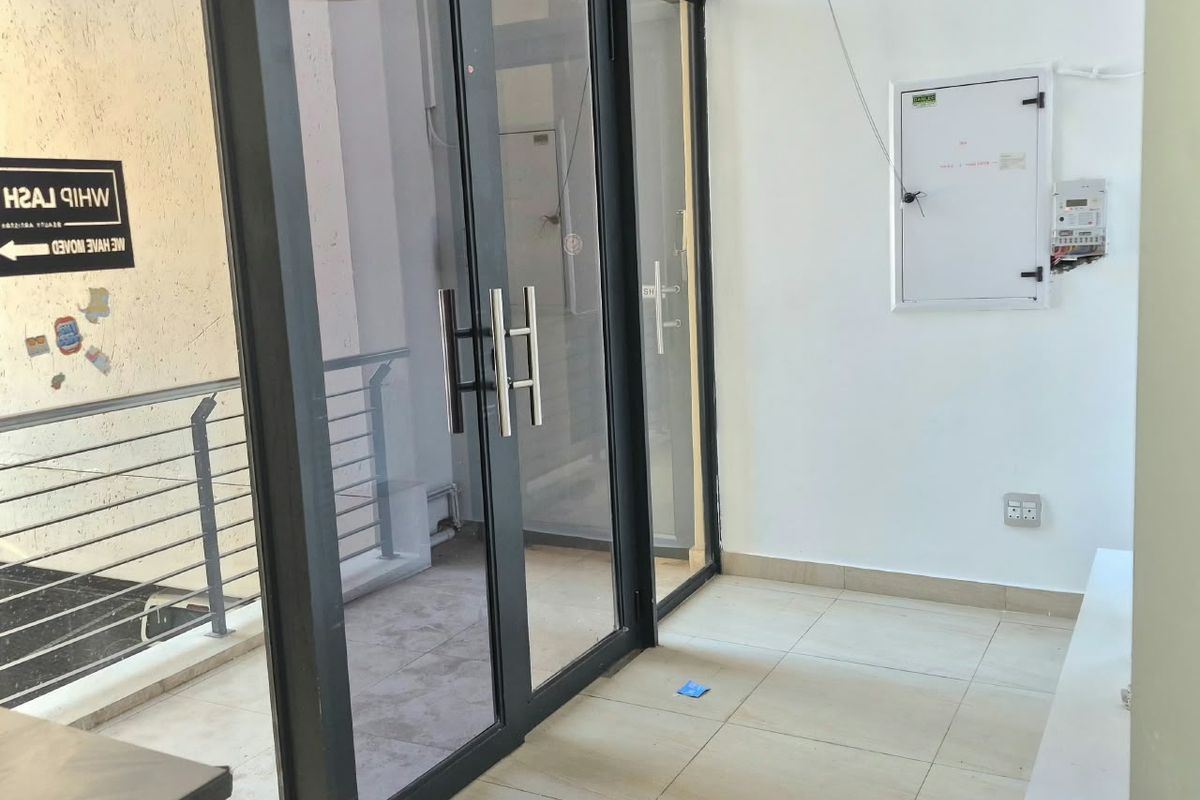 A bright office entrance area featuring modern aluminium framed glass double doors, neutral tiled flooring, and a wall-mounted electrical distribution board.