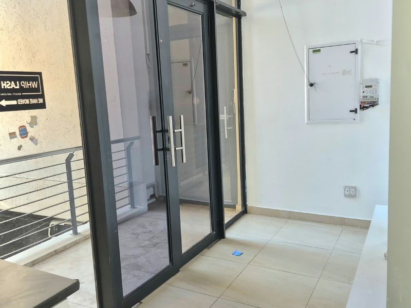 A bright office entrance area featuring modern aluminium framed glass double doors, neutral tiled flooring, and a wall-mounted electrical distribution board.