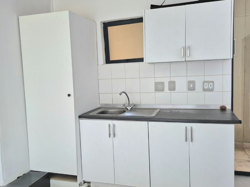 A compact kitchenette featuring white built-in cabinetry, a stainless steel sink, tiled backsplash, and dark laminate countertops in a clean, functional office space.