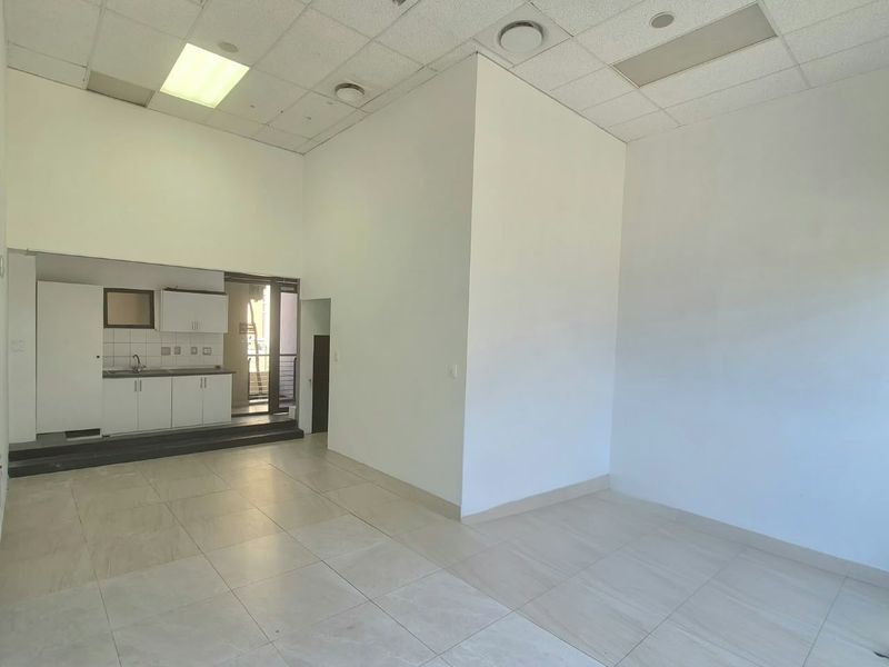 An open-plan office space featuring tiled flooring, a suspended ceiling with recessed lighting, and a compact kitchenette area with built-in cabinetry.
