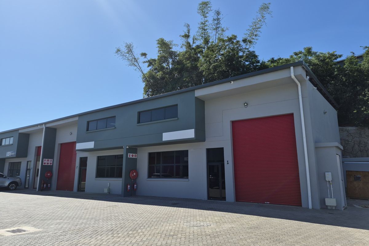 Modern industrial warehouse unit featuring a large red roller shutter door, paved exterior frontage, and contemporary grey and dark green facade finishes.