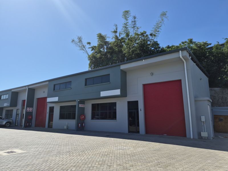 Modern industrial warehouse unit featuring a large red roller shutter door, paved exterior frontage, and contemporary grey and dark green facade finishes.
