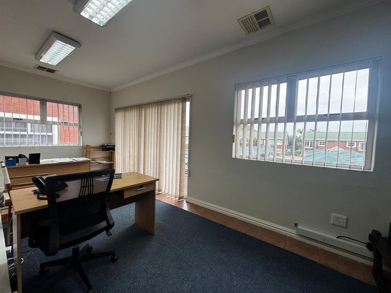 A functional office space featuring carpeted flooring, vertical blinds, security bars on windows, and integrated ceiling lighting with air conditioning ventilation.