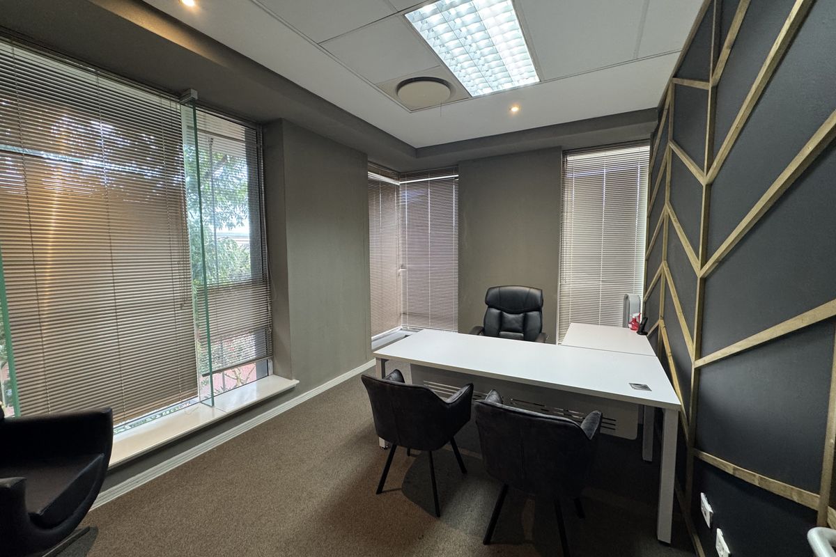 A modern professional office space featuring large windows with venetian blinds, decorative wall paneling, carpeted flooring, and recessed ceiling lighting.
