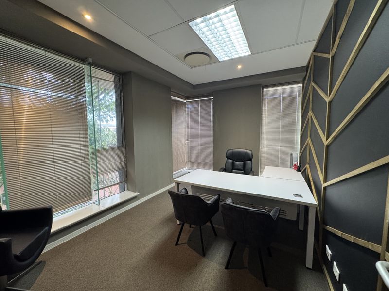 A modern professional office space featuring large windows with venetian blinds, decorative wall paneling, carpeted flooring, and recessed ceiling lighting.