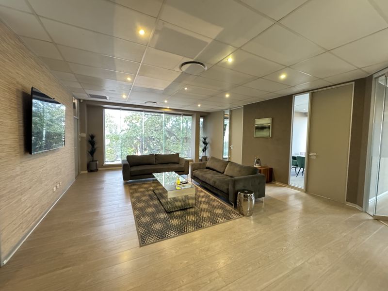 A modern office lounge area featuring a wood-panelled feature wall, suspended ceiling with recessed lighting, and large windows providing natural light.