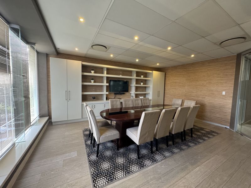 A professional office boardroom featuring wood-look wall paneling, a large built-in shelving unit, suspended ceiling with recessed lighting, and expansive windows with blinds.