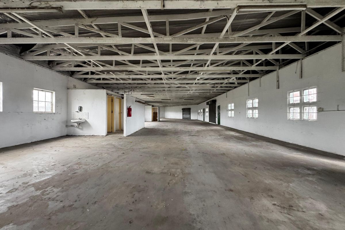 Spacious industrial open-plan warehouse area featuring exposed steel roof trusses, concrete flooring, and multiple windows providing natural light.