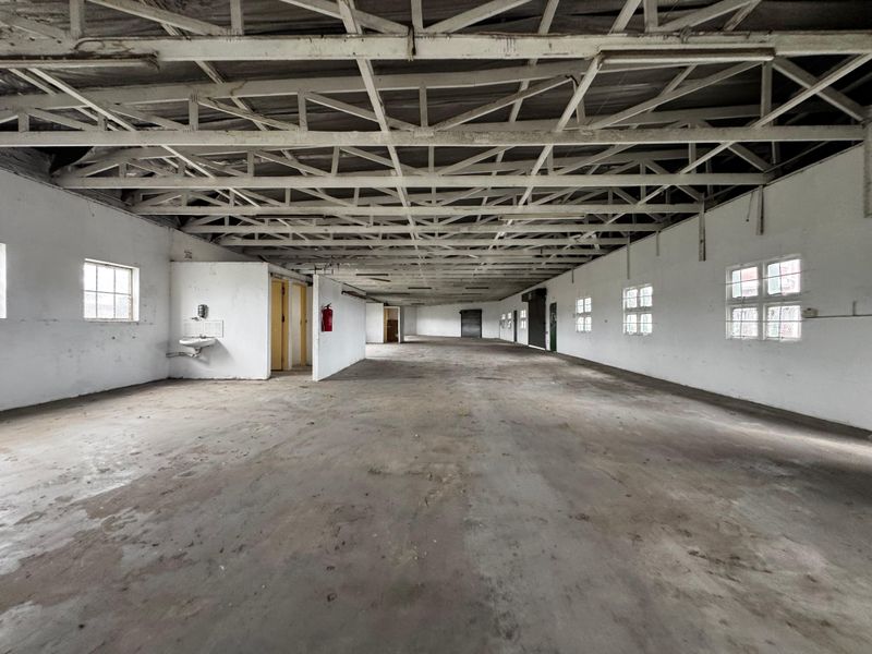 Spacious industrial open-plan warehouse area featuring exposed steel roof trusses, concrete flooring, and multiple windows providing natural light.